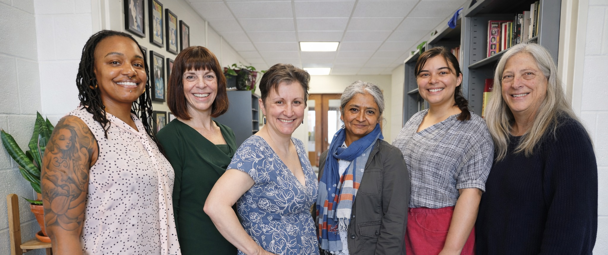 Six women standing together in a hallway, smiling warmly. Shelves with books are visible in the background, conveying a professional setting.